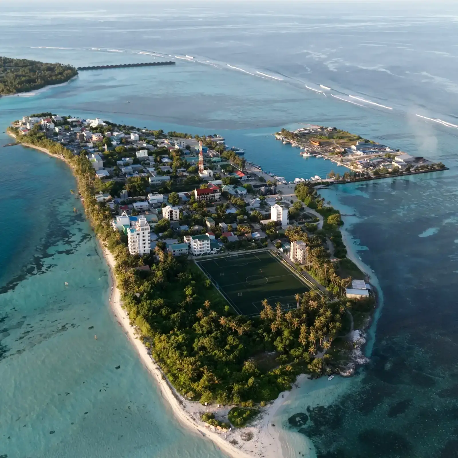 Aerial view of Dhiffushi in early morning light, with the village, harbour and football ground inside a wide turquoise lagoon